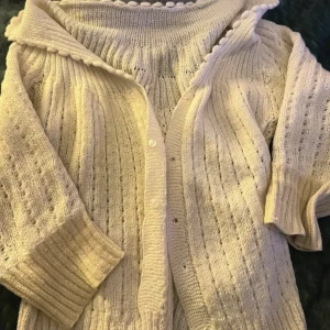 Vit stickad kofta med knappar - Säljer en vit stickad kofta med ribbad struktur och dekorativa hål. Koftan har trekvartsärmar, rundad krage med små bollar och knäppning framtill. Perfekt för dig som gillar mysiga och stilrena plagg med fina detaljer. Andra bilden är mer rättvis på färgen på koftan men på första kan man se att slutet av ärmarna är lite mer åt de gula hållet