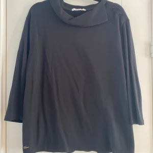 Svart polotröja från Lacoste - Svart polotröja från Lacoste i 100% bomull med cool krage och knappar på axeln. Tröjan har trekvartsärm och den klassiska krokodilloggan nertill. Perfekt för dig som gillar stilren och enkel design. Mått Över bröstet: ca 56 cm Längd från axeln och ner: ca 62 cm  100% bomull  Kommer från ett djur och rökfritt hem.