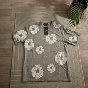 Grå t-shirt med vita blommor från  Denim Tears - Säljer en grå t-shirt från Denim Tears med stora vita blommönster över hela plagget. T-shirten har rund hals och korta ärmar, tillverkad i 100% bomull. Märket syns tydligt i nacken och på etiketten. Perfekt för dig som gillar unik streetwear.