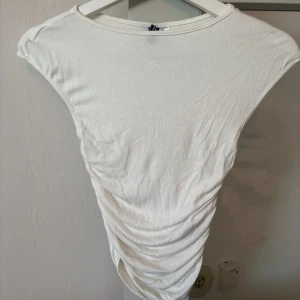 Vit ärmlös croppad topp - Säljer en vit ärmlös topp med croppad passform och snygga rynkade detaljer längs sidorna. Toppen har rund halsringning och är tillverkad i mjuk bomull, perfekt för en chill och stilren look.