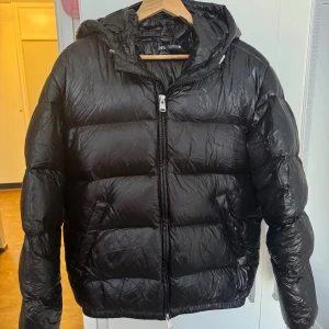 Svarta virala pufferjacka från Zara  - Säljer en svart pufferjacka från Zara med glansig finish och huva. Jackan har dragkedja framtill, två sidofickor och quiltad design. Perfekt för kalla dagar när du vill hålla stilen och värmen.