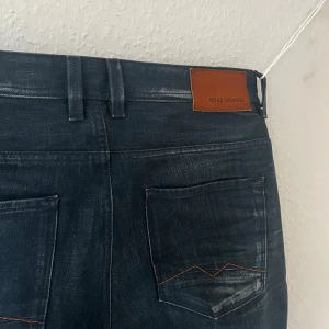 Boss Orange mörkblå jeans - Snygga mörkblå jeans från Boss Orange med klassisk femficksdesign och orangea detaljer på knappar och etikett. Jeansen har en rak passform och snygga slitningar. De bilder som visar hela byxan är lite missvisande, byxorna har färgen som de första två bilder visar.