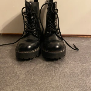 Svarta boots med snörning och grov sula - Säljer ett par svarta boots med snörning och grov platt sula. Skorna har rund tå, metallöglor och är tillverkade i skinn. Perfekta för dig som gillar en tuff och stilren look. Passar till både jeans och kjol.Perfektta höst skor 🍁