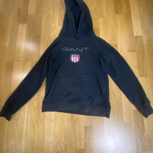 Svart hoodie från GANT med logga - Svart hoodie från GANT med broderad logga och sköld på bröstet. Klassisk känguruficka framtill och huva. Perfekt för en chill och sportig stil.