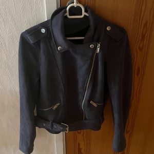 Svart mocka bikerjacka med bälte - Snygg mörkblå bikerjacka i mocka med asymmetrisk dragkedja, silverfärgade detaljer och bälte i midjan. Jackan har långa ärmar och två dragkedjefickor framtill. Perfekt för dig som vill ha en edgy och stilren look.🤩😁