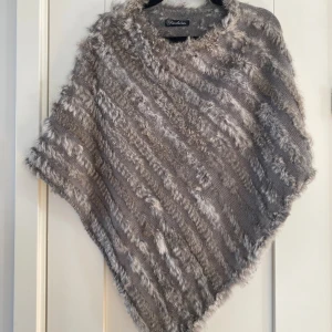Grå poncho med kaninpäls från Fashion - Säljer en fluffig och mysig grå poncho från Fashion, tillverkad av 90% kaninpäls och 10% akryl. Den har ett snyggt randigt mönster och är perfekt för att hålla sig varm under kyliga dagar. Ponchon har en unik form med korta ärmar och mjuk pälsstruktur.