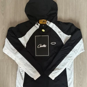 Corteiz - Svart och vit vindjacka från Corteiz med huva och dragkedja. Jackan har kontrasterande vita paneler längs ärmar och sidor, samt Corteiz-logga på bröstet och ryggen. Tillverkad i lätt polyester, perfekt för streetwear-stil. Långärmad och med elastiska muddar. Helt oanvänd med prislapp på!