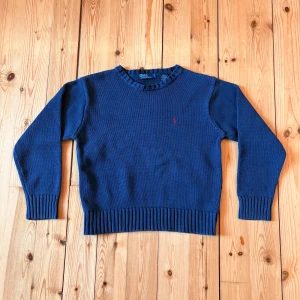 Marinblå stickad tröja Polo Ralph Lauren - Säljer en marinblå stickad tröja från Polo Ralph Lauren i 100% bomull. Tröjan har rund halsringning, ribbade muddar och nederkant samt en liten röd broderad logga på bröstet. Perfekt för lager-på-lager under kyliga dagar.