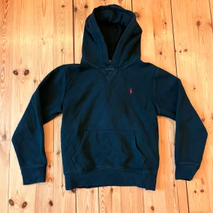 Svart hoodie från Polo Ralph Lauren - Svart hoodie från Polo Ralph Lauren med klassisk röd broderad logga på bröstet. Tröjan har huva, känguruficka och ribbade muddar. Perfekt för dig som gillar stilren och enkel design med en touch av lyx.