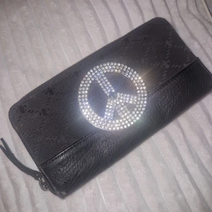 Plånbok  - Svart plånbok från Friis & Company i skinn med dragkedja runtom och flera fack för kort och mynt. Framsidan har ett stort peace-märke i glittrande stenar som ger en cool vibe. Perfekt storlek för att få plats med allt du behöver. Finns lite små defekter