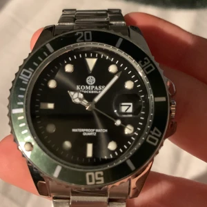 Kompass OceanXplorer Silver Green Black - Snygg Kompass OceanXplorer klocka med robust silverfärgad länk i metall, svart urtavla och grön bezel. Klockan har tydliga index, datumvisning och quartz-urverk. Perfekt för dig som gillar stilren design med sportig känsla.