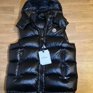 Svart dunväst från Moncler - Snygg svart dunväst från Moncler med glansig finish, huva och dragkedja framtill. Västen har två fickor med dragkedja och ikonisk Moncler-logga på bröstet. Insidan har ett coolt serietidningsmönster och märkets etikett. Perfekt för lager-på-lager-stil.
