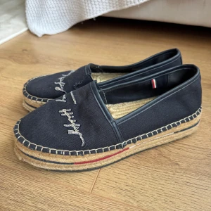 Marinblå  espadrillos Tommy Hilfiger - Snygga marinblå espadrillos från Tommy Hilfiger med broderad logga i silver på ovansidan. Skorna har en rund tå, platt sula i jute med dekorativa sömmar och en diskret röd detalj på sidan. Ovandelen är i bomullstyg och insidan har en liten Hilfiger-etikett.
