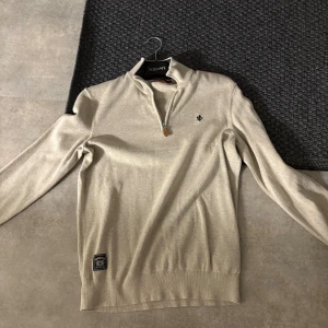 Beige halvzip tröja från Morris - Snygg beige långärmad tröja från Morris med half zip och liten broderad logga på bröstet. Tröjan har ribbade muddar och en patch nertill. Perfekt för dig som gillar stilren och enkel design.