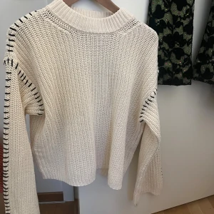 Beige stickad tröja med svarta detaljer - Snygg beige stickad tröja med grov struktur och svarta sömdetaljer längs ärmarna. Tröjan har en hög ribbad hals och långa ärmar, perfekt för att hålla dig varm och stilren under kyliga dagar.