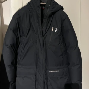 Svart dunjacka Peak Performance - Svart dunjacka från Peak Performance med huva och dragkedja framtill. Jackan har flera fickor, diskret logga på bröstet och en på fickan. Perfekt för kalla dagar. Modell: style S W D parka. Näsan oanvänd!