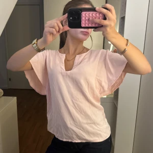 Ljusrosa blus med v-ringning - Säljer en ljusrosa blus med lös passform och v-ringning. Blusen har korta, vida ärmar och är tillverkad i ett tunt, mjukt tyg som känns luftigt. Perfekt för dig som gillar en enkel men söt stil.. Märke SAINT TROPEZ