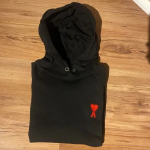 Svart hoodie från Ami Paris med rött hjärta - Svart hoodie från Ami Paris med ikoniskt rött hjärta och A broderat på bröstet. Klassisk huva med snörhål, mjuk bomullskvalitet och ribbade muddar. Perfekt för en chill och stilren look.