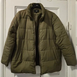 Olivgrön pufferjacka från Zizzi - Säljer en olivgrön pufferjacka från Zizzi med quiltad design och hög krage. Jackan har dragkedja framtill, två sidofickor och en loose passform. Perfekt för kyliga dagar och enkel att matcha med olika outfits.