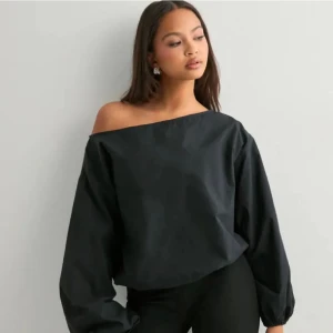 Svart offshoulder blus med lång ärm - Säljer en svart blus med offshoulder-design och långa, puffiga ärmar. Toppen har en lös passform och är tillverkad i ett slätt material som ger en stilren look. Perfekt för dig som gillar minimalistisk stil och vill ha något som sticker ut med sin asymmetriska halsringning. Den är i nytt skick 