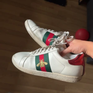  Ace sneakers med bi-detalj -  Ace sneakers i vitt skinn med klassiska gröna och röda ränder på sidan och broderad guldfärgad bi. Röd hälpanel med textur och snörning framtill. Rund tå och platt sula med -logga undertill. Perfekt för dig som gillar lyxiga streetwear-vibes.