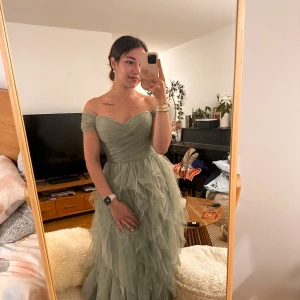 Ljusgrön offshoulder balklänning - Superfin ljusgrön långklänning med offshoulder och hjärtformad urringning. Klänningen har flera lager volangigt tyg som ger en drömmig känsla och markerad midja. Perfekt för bal eller speciella tillfällen. Materialet är tunt och skirt, troligtvis polyester eller liknande.