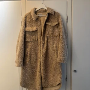 Beige teddyjacka från NA-KD - Säljer en lång beige teddyjacka från NA-KD med två bröstfickor samt på sidorna och knappar framtill. 