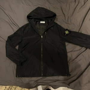 Svart vindjacka från Stone Island - Svart vindjacka från Stone Island med ikonisk patch på ärmen. Jackan har huva, hel dragkedja framtill och är tillverkad i lätt polyester. Perfekt för dig som gillar streetwear och vill ha en stilren look.