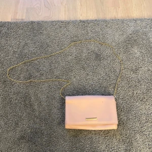 Ljusrosa väska med axelband kedja - Snygg ljusrosa kuvertväska med guldfärgad kedja som axelrem. Insidan har fack för kort och en dragkedja för småsaker. Har en fläck på utsidan