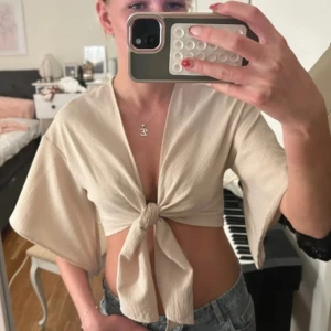 Beige croppad knytblus med vida ärmar - Snygg croppad blus i beige med knyt framtill och vida, korta ärmar. Toppen har en lätt textur i tyget och ger en chill och trendig vibe. Perfekt att styla med jeans eller kjol för en cool look.