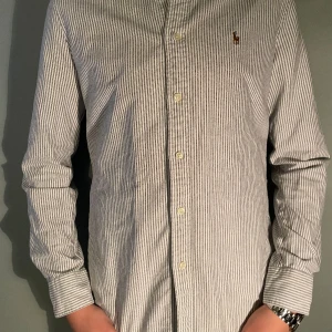 Randig skjorta från Ralph Lauren - Säljer en klassisk skjorta från Ralph Lauren med smala vertikala ränder i vitt och grått. Modellen är 177-178cm lång, skjortan har lång ärm, knappar framtill och den ikoniska broderade loggan på bröstet. Tillverkad i bomull för en skön och luftig känsla.