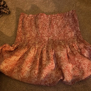 Rosa kortkjol från Gina Tricot - Supersöt kortkjol från Gina Tricot i ljusrosa med röd paisley-inspirerat mönster. Kjolen har smockad midja som ger en skön och flexibel passform, samt volangig nederdel. Perfekt för dig som gillar boho-vibe och vill ha något luftigt och snyggt.