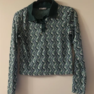 Mönstrad pikétröja från Zara - Snygg pikétröja från Zara i storlek S med retroinspirerat geometriskt mönster i grönt, blått och beige. Tröjan har klassisk krage, knappar framtill och långa ärmar. Perfekt för dig som gillar unika prints och vill sticka ut lite extra.