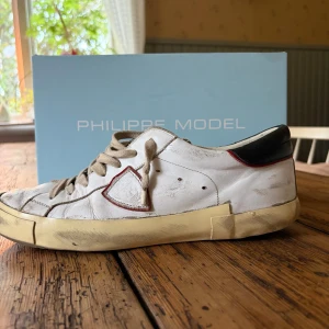 Vita Philippe Model sneakers i skinn - Säljer ett par vita Philippe Model sneakers i skinn med beige sula och svarta detaljer vid hälen. Skorna har snörning och en röd kant runt loggan på sidan. Klassisk låg modell med rund tå och vintage-inspirerad look. Slitingarna ser ni på bilderna, gäller endast på högra skon! Dom behöver kärlek!🥰Passar 44/45