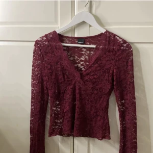 Vinröd spetsblus från Gina Tricot - Vinröd långärmad blus från Gina Tricot i transparent spets med blommigt mönster. V-ringad framtill och figurnära passform. Perfekt för dig som gillar romantiska detaljer och vill sticka ut med en djup vinröd ton. Blusen har 3 jättesmå hål som inte märks om man inte kollar super noga. 