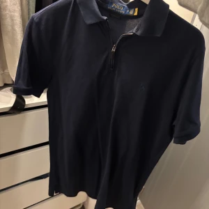 Marinblå pikétröja Polo Ralph Lauren - Snygg marinblå pikétröja från Polo Ralph Lauren med klassisk krage och half zip-dragkedja. Tröjan har korta ärmar och är i en slim fit-modell. Tillverkad i bomull med stretch för extra komfort. Perfekt för en clean och stilren look.