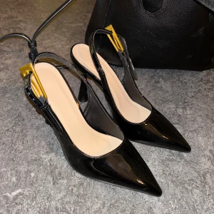 Svarta lackpumps med gulddetalj - Säljer ett par svarta pumps i lack med spetsig tå och öppen häl. Skorna har en elegant hög klack och snygga guldfärgade spännen vid remmen. Perfekta för dig som vill sticka ut med en stilren och trendig look.