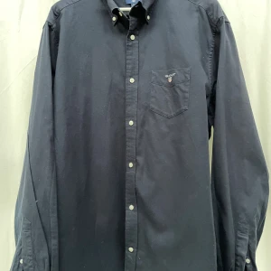 Mörkblå långärmad skjorta från GANT XL - Mörkblå skjorta från GANT i storlek XL med klassisk button-down krage och bröstficka med broderad logga. Skjortan har vita knappar framtill och på ärmsluten. Tillverkad i mjuk bomull, perfekt för dig som gillar stilren och enkel design. Nypris 1299kr