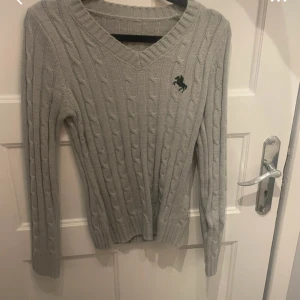 Grå stickad tröja från Shein - Snygg grå stickad tröja från Shein i storlek M. Tröjan har ett klassiskt kabelstickat mönster, v-ringad hals och långärmade ärmar. På bröstet finns en liten svart broderad häst. Perfekt för dig som gillar enkel och stilren look.