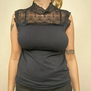 Svart ärmlös topp med spetsdetalj - Snygg svart ärmlös topp med hög hals och transparent spets upptill. Spetsen har ett vågigt mönster och täcker axlarna samt halsen för en cool look. Resten av toppen är i mjukt tyg som sitter bekvämt mot kroppen. Perfekt för dig som gillar stilrena detaljer. Men passar en M