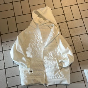 Vit cardigan jacka  - Säljer en stilren vit cardigan jacka med quiltad front och dragkedja. Tröjan har huva, långa ärmar och två fickor med dragkedja. Perfekt för dig som gillar en clean look och vill ha något bekvämt och snyggt till vardags. Priset är inte hugget i sten!