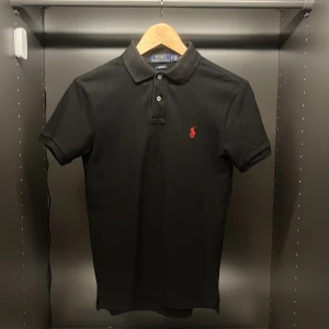 Svart pikétröja från Ralph Lauren - Klassisk svart pikétröja från Polo Ralph Lauren i slim fit-modell. Tröjan har korta ärmar, krage och knäppning framtill samt den ikoniska röda loggan broderad på bröstet. Perfekt för en clean och stilren look. Storlek S.