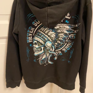 Svart hoodie Rock Rebel med tryck - Svart hoodie från Rock Rebel med dragkedja, huva och snörning. Stor tryckt design på ryggen med dödskalle och fjädrar i blått och vitt, samt broderat framtill!🥰🥰Inga defekter!