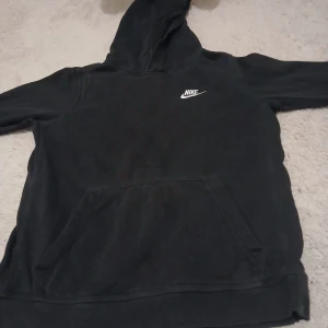 Svart hoodie från Nike med huva - Svart hoodie från Nike med klassisk logga broderad i vitt på bröstet. Tröjan har en stor magficka, huva och långa ärmar. Perfekt för chill eller träning, enkel och stilren design som funkar till det mesta.