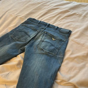 Blå jeans från Emporio Armani - Snygga blå jeans från Emporio Armani med klassisk femficksdesign och ikonisk örnlogga på bakfickan. Jeansen har en rak passform och subtila slitningar för en cool look. Perfekta för dig som vill ha stilrena och bekväma jeans med exklusiv känsla.