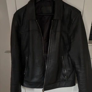 Svart jacka Bolongaro Trevor Strl L (passar M) - Snygg svart jacka i konst-skinn från Bolongaro Trevor i storlek L men passar även M. Jackan har klassisk krage, raka linjer och dragkedjor både framtill och vid fickorna. Tillverkad i konst skinn med slät finish och helsvart foder. Perfekt för dig som gillar stilren och tidlös look.