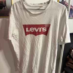 Vit Levi's t-shirt med röd logga - Vit t-shirt från Levi's med klassisk röd logga framtill. T-shirten har rund halsringning och korta ärmar. Tillverkad i mjuk bomull för en skön känsla. Perfekt för en avslappnad och stilren look.