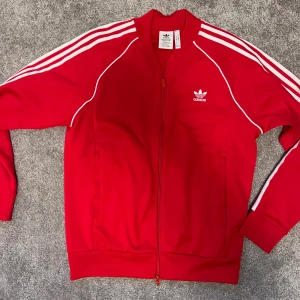Röd Adidas track hoodie med vita ränder - Röd Adidas track jacket med klassiska vita ränder längs ärmarna och broderad logga på bröstet. Jackan har dragkedja framtill, ribbade muddar och vita detaljer längs sömmarna. Tillverkad i polyester för en sportig vibe.