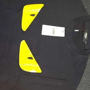 Svart Fendi t-shirt med gula detaljer - Svart t-shirt från Fendi med två stora gula ögonliknande detaljer framtill. Klassisk rund halsringning och korta ärmar. Tillverkad i mjuk bomull för en skön känsla. Perfekt för dig som vill sticka ut med en unik designerstil.