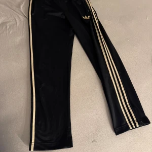 Svarta Adidas byxor med guldiga ränder - Svarta Adidas byxor med guldiga ränder längs sidorna och klassisk treklöverslogga. Byxorna har dragkedjor vid fickorna och bensluten, samt resår i midjan. Tillverkade i ett glansigt syntetmaterial som ger en sportig vibe. Dessa säljs inte längre 